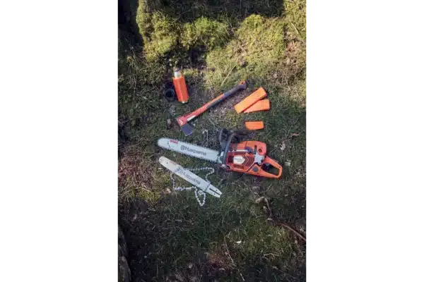 Lesnícke vybavenie Husqvarna na zemi, vrátane motorovej píly, sekery, termosky a dvoch oranžových plastových klinov.