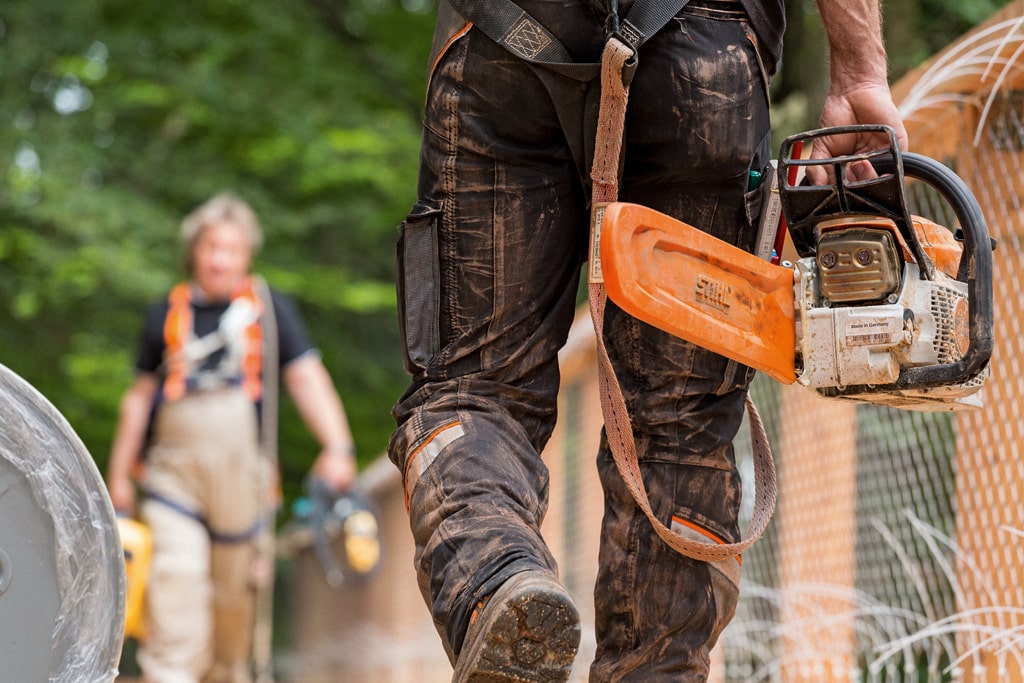 Drevorubač v zablatených protiporezových nohaviciach kráča a v pravej ruke nesie motorovú pílu STIHL.